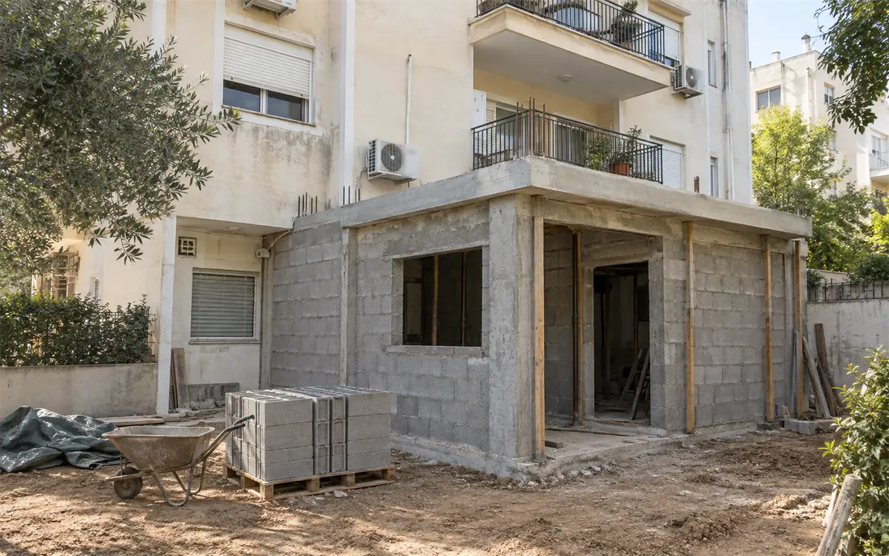 תמונה להמחשה: חצר אחורית של בניין מגורים ותיק, ובקומת הקרקע תוספת חדר חדשה שנבנית לתוך החצר: שלד בטון עם עמודים ותקרה, מילוי בבלוקים אפורים שעוד לא טויחו, פתח חלון ופתח דלת ריקים, קוצי ברזל בולטים מהתקרה ותמוכות עץ, ומשטח בלוקים ומריצה על האדמה
