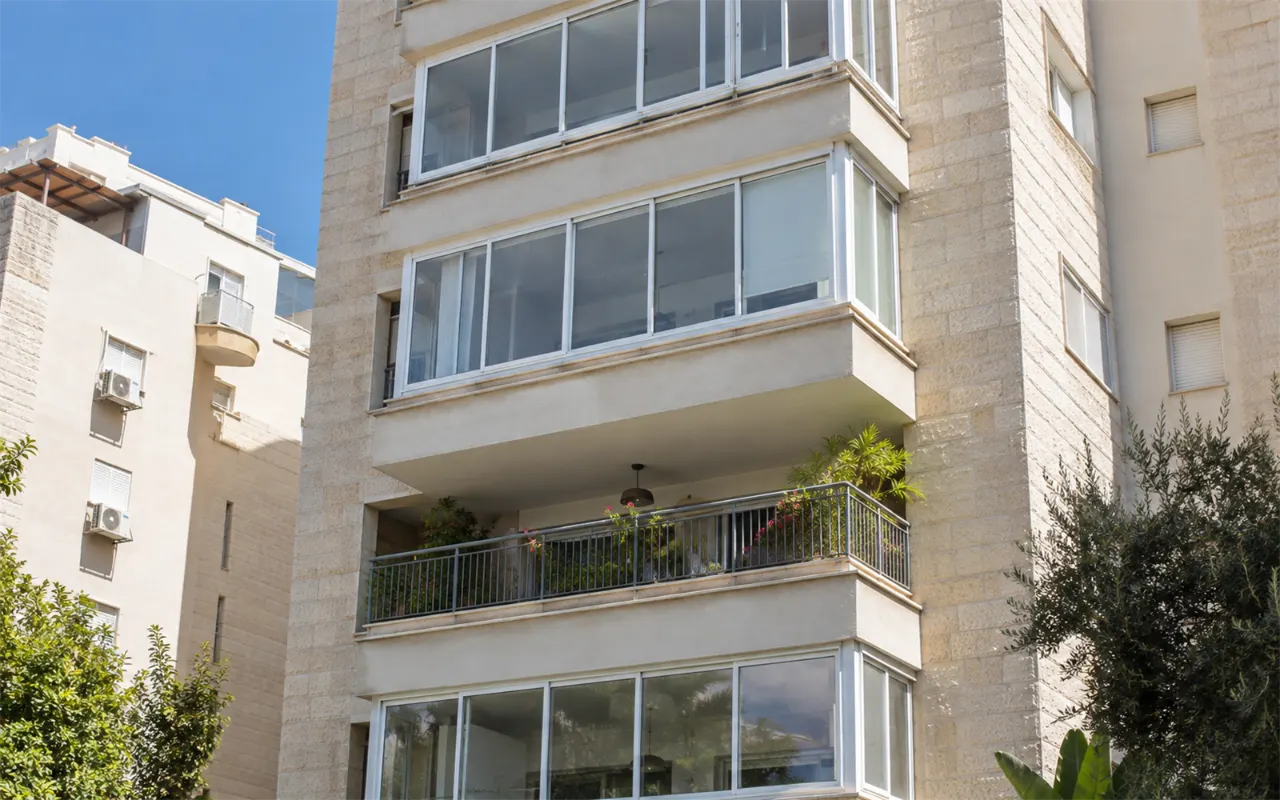 תמונה להמחשה: חזית בניין מגורים משנות התשעים עם מרפסות זו מעל זו: כמה מהן סגורות באופן אחיד באותה מערכת חלונות הזזה מאלומיניום לבן, ומרפסת אחת עדיין פתוחה עם מעקה ועציצים, על רקע חיפוי אבן בהיר