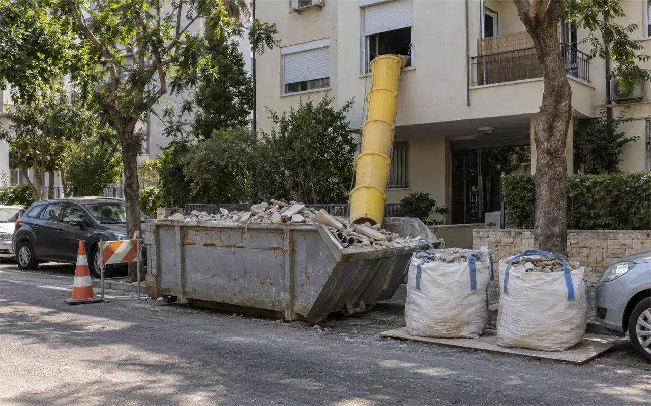 תמונה להמחשה: רחוב מול בניין מגורים בזמן שיפוץ: מכולת פסולת מפלדה בשולי הכביש מלאה בשברי אריחים, בטון וצנרת ישנה, שרוול פסולת צהוב שיורד מחלון בקומה הראשונה אל המכולה, שני שקי בלה עם פסולת על משטח הגנה לידה, קונוס וגדר סימון קטנה